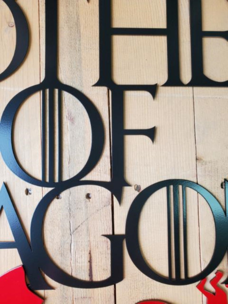 May include: Black metal wall art with the words "House of the Dragon" in a stylized font. The letters are cut out and mounted on a wood background.