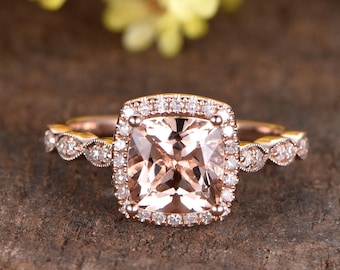 May include: A rose gold engagement ring with a square-shaped morganite center stone surrounded by a halo of smaller diamonds. The band is adorned with a delicate, scalloped design and smaller diamonds.