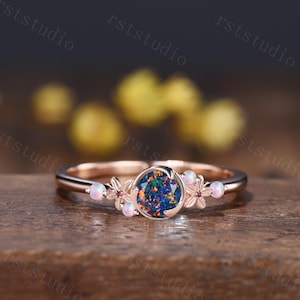 May include: A rose gold ring featuring a round, dark, iridescent opal center stone. The band is accented with small, delicate metal flowers and four tiny, round, white opals. The design is symmetrical, with the floral elements flanking the central stone.