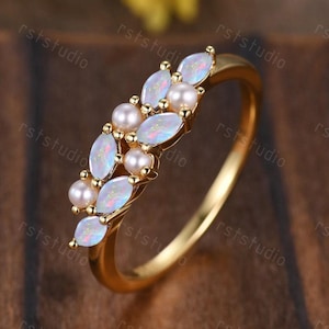 May include: A gold ring with a delicate design featuring white opal stones and small pearls. The ring is set on a wooden surface.