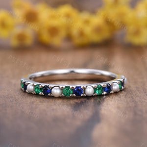 May include: A silver band ring featuring alternating green and blue gemstones, interspersed with small white pearls. The ring is set on a wooden surface, with a blurred yellow background.
