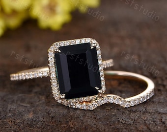 May include: A black emerald cut engagement ring with a halo of small diamonds set in yellow gold. The ring is paired with a matching wedding band with a curved design.