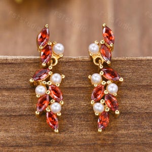 May include: A pair of gold-toned earrings featuring a leaf-like design. Each earring has red, marquise-cut gemstones and small, round, white pearls. The earrings are set against a wooden background.