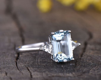 May include: A silver ring with a large, light blue emerald-cut aquamarine gemstone and two smaller, triangular diamonds on either side.