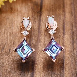 May include: A pair of rose gold dangle earrings with a square-shaped blue gemstone, a small round white gemstone, and a teardrop-shaped white opal.