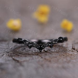 May include: A black ring with a curved design, featuring leaf and gemstone accents. The ring has a V-shape with small black gemstones and leaf-like details. The ring is set against a blurred background.