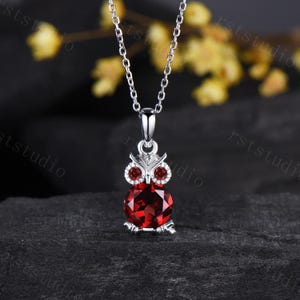 May include: A silver-toned necklace featuring an owl-shaped pendant. The owl has two small red circular stones for eyes and a large, round, faceted red stone forming its body. The pendant hangs from a delicate silver-toned chain link necklace.