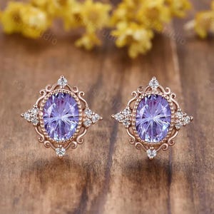 May include: A pair of rose gold stud earrings. Each earring features an oval, light purple gemstone surrounded by small white gemstones and a decorative filigree frame. The earrings are set against a wooden background.