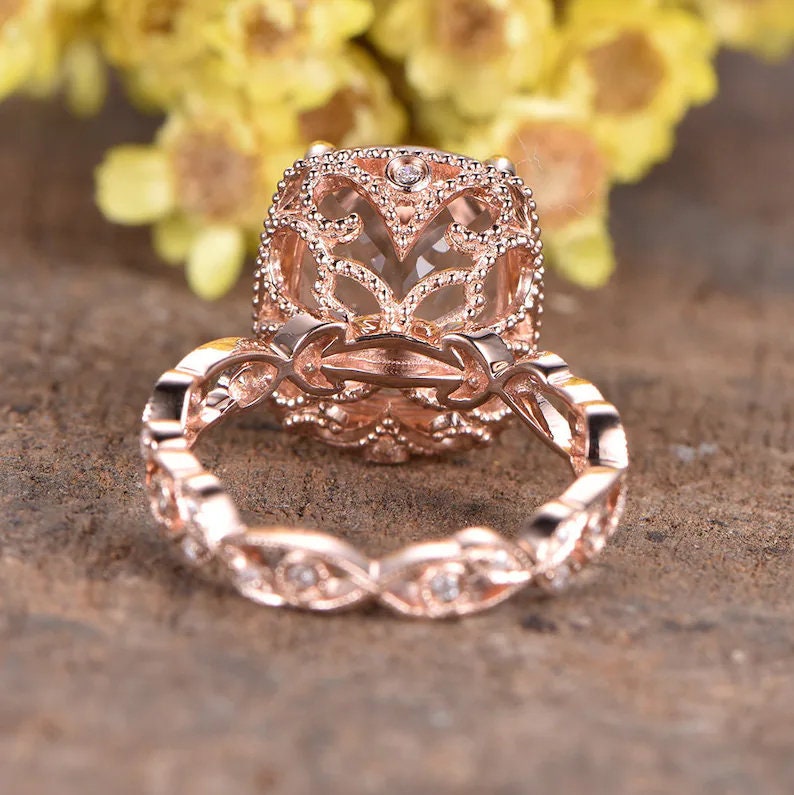 May include: Rose gold engagement ring featuring a large, clear, oval-shaped gemstone set in a delicate, filigree design. The band is adorned with smaller diamonds.
