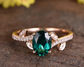 May include: A rose gold engagement ring with a large oval emerald green gemstone surrounded by smaller diamonds. The ring features a delicate leaf design on each side of the center stone.