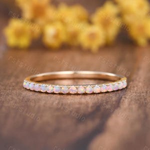 May include: A delicate gold ring featuring a continuous row of small, round opals. The opals are a soft, milky white color, set closely together along the band. The ring is displayed on a warm, brown surface, with blurred yellow flowers in the background.