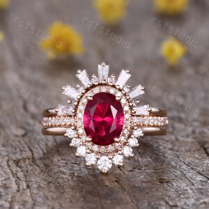 Puede incluir: Un anillo de compromiso de oro rosa con un gran rubí ovalado rodeado de diamantes más pequeños. El anillo tiene un diseño delicado de inspiración vintage con un halo de diamantes y una banda de diamantes más pequeños.