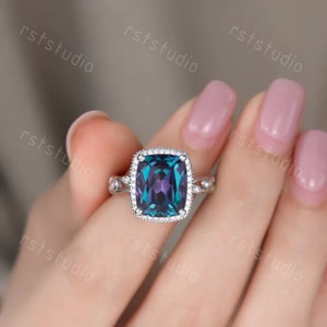 May include: A silver ring featuring a large, square-shaped, blue and purple gemstone surrounded by smaller, clear gemstones. The ring is set on a white band with a delicate design.