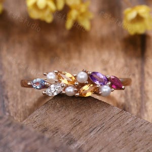 May include: A gold-toned ring featuring a cluster of gemstones and pearls. The ring showcases a variety of colorful stones, including blue, white, yellow, purple, and pink, interspersed with small, round pearls. The gemstones are set in a leaf-like arrangement.