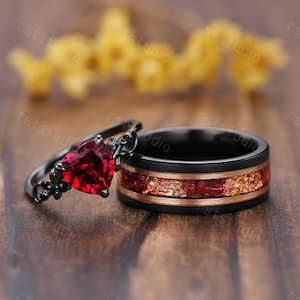 May include: A pair of wedding rings. One ring features a heart-shaped, deep red gemstone and a black band with floral accents. The other ring has a black band with a copper-colored inlay and a red center.
