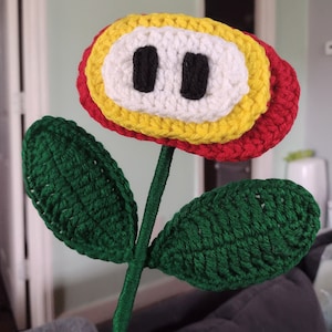 May include: A crocheted flower with red, yellow, and white petals and green leaves. The flower has a white center with two black circles.