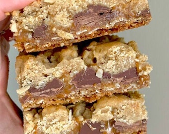 Peut inclure: Trois barres de dessert superposées avec une garniture dorée et friable et une couche de chocolat. Les barres ont une base de caramel et des morceaux de chocolat visibles. Image en gros plan.