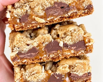 May include: A stack of three square oatmeal cookie bars featuring a crumbly golden-brown texture, visible chunks of dark chocolate, and a thick, gooey layer of amber-colored caramel filling sandwiched between the baked oat layers.