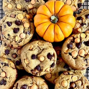 May include: A collection of round, golden-brown cookies studded with dark chocolate chips and orange butterscotch chips, arranged around a single, small, vibrant orange pumpkin with a green stem, all resting on a black wire cooling rack.