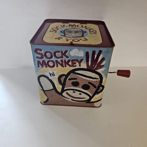 May include: A vintage-style Jack-in-the-box toy with a red crank. The box is a square shape with a maroon lid and blue sides. The box features a sock monkey design with the words "Sock Monkey" and "hi".
