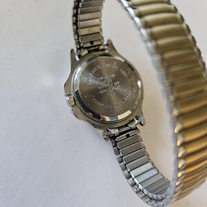 May include: A silver-toned wristwatch with a stretch band. The watch face is not visible, but the back shows text including "BASE METAL BEZEL" and "STAINLESS STEEL BACK". The band is a mix of silver and gold tones.