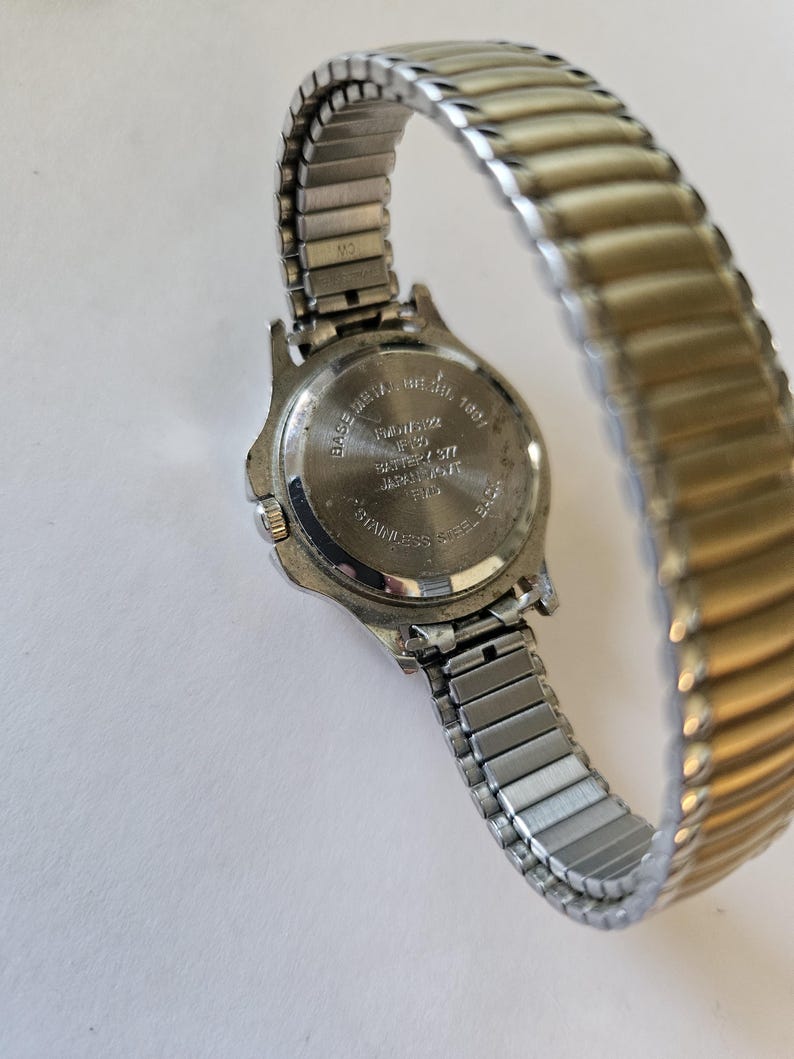 May include: A silver-toned wristwatch with a stretch band. The watch face is not visible, but the back shows text including "BASE METAL BEZEL" and "STAINLESS STEEL BACK". The band is a mix of silver and gold tones.