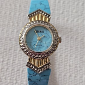 May include: A turquoise-colored watch with a gold-colored bezel and band accents. The watch face has the word "ITANO" and "QUARTZ" printed on it. The watch band is made of turquoise-colored material.