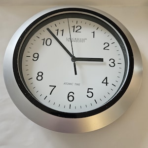 May include: A round wall clock with a silver frame and a white face featuring black Arabic numerals. The clock displays the text La Crosse Technology and Atomic Time. It has three black hands indicating the time, with the hour hand pointing toward three, the minute hand toward ten, and the second hand near twelve.