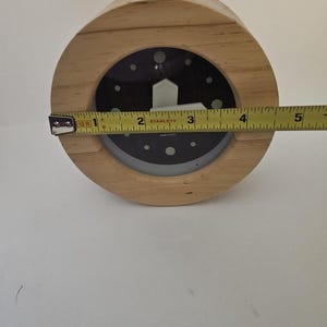 Puede incluir: Un reloj de madera con esfera negra y manecillas blancas. El reloj tiene aproximadamente 10 cm de di&aacute;metro. El reloj es redondo y tiene un marco de madera marr&oacute;n claro.