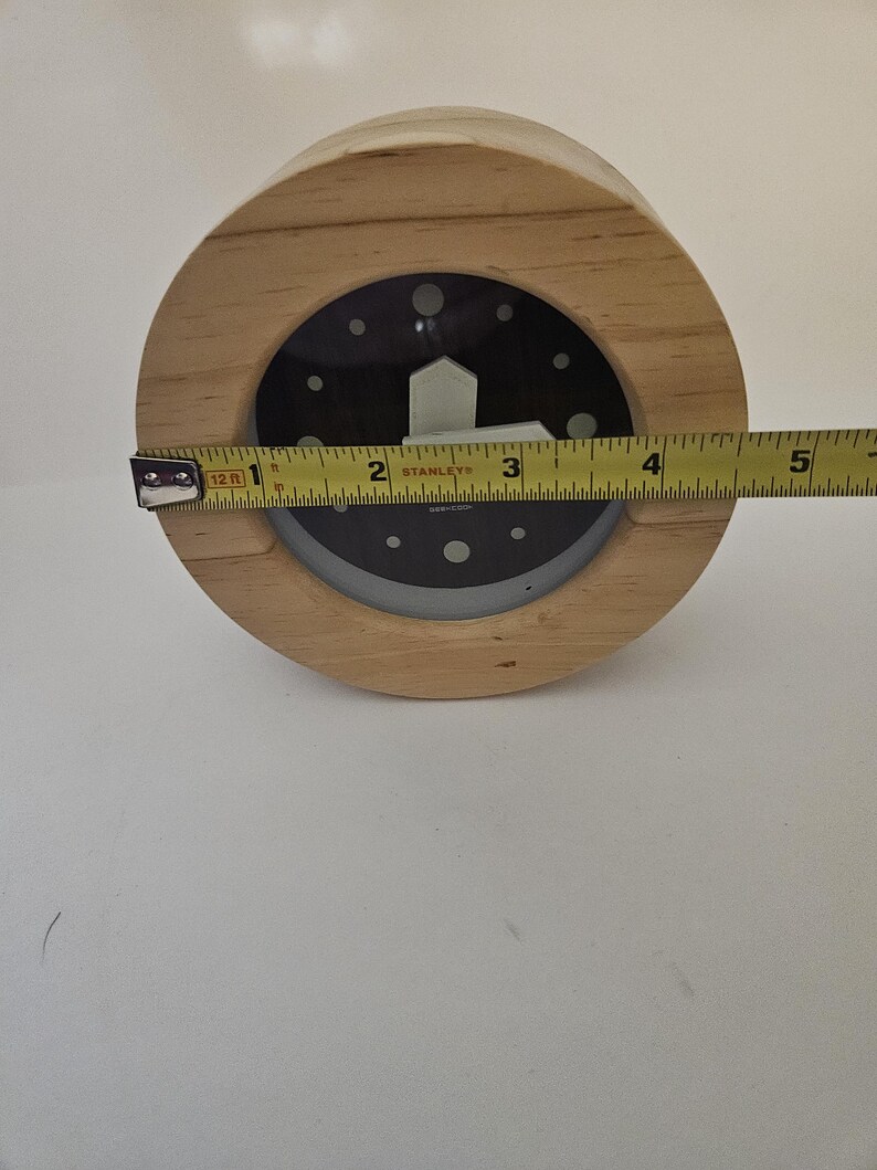 Puede incluir: Un reloj de madera con esfera negra y manecillas blancas. El reloj tiene aproximadamente 10 cm de di&aacute;metro. El reloj es redondo y tiene un marco de madera marr&oacute;n claro.