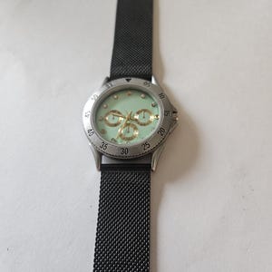 May include: A wristwatch with a black mesh metal strap and a silver-tone circular case. The watch face is a pale mint green color featuring gold-tone hands, three small sub-dials, and small circular hour markers. The bezel is silver with black numerals.