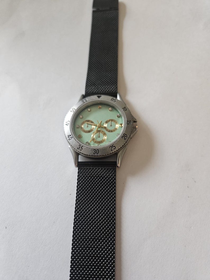 May include: A wristwatch with a black mesh metal strap and a silver-tone circular case. The watch face is a pale mint green color featuring gold-tone hands, three small sub-dials, and small circular hour markers. The bezel is silver with black numerals.