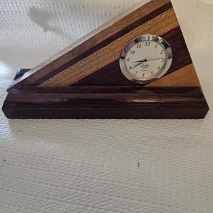 May include: A wooden desk clock with a triangular design. The clock features a silver-colored clock face with black numbers and hands. The wooden base is a dark brown color with lighter brown stripes.