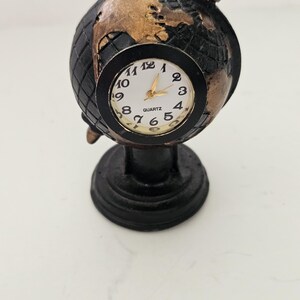 May include: A black and gold globe-shaped clock with a white clock face. The clock has gold hands and a black base. The clock face reads "QUARTZ". The globe has a textured surface and is mounted on a round base.
