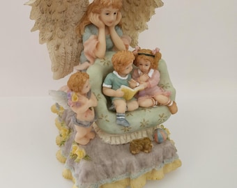 Puede incluir: Figurilla detallada que representa un ángel con grandes alas, observando a tres niños leyendo un libro. Los niños están sentados en una silla con estampado floral. Un pequeño querubín está cerca. La base tiene una falda con volantes y ribete amarillo.