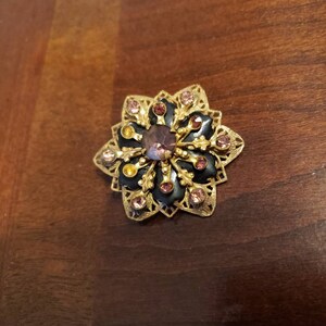 May include: A gold-toned brooch with a black enamel center and pink and yellow rhinestones. The brooch is shaped like a flower with intricate details.