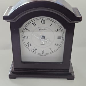 May include: A dark brown wooden mantel clock with an arched top and a rectangular base. The white clock face features black Roman numerals, black hands, and the text WALLACE and QUARTZ printed in black. The bottom edge reads MADE IN CHINA.