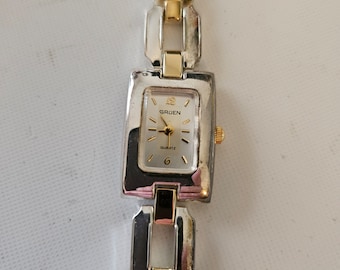 May include: A silver and gold two-tone watch with a rectangular face. The watch face is white with black markings and the word "GRUJEN" and "QUARTZ" printed on it.