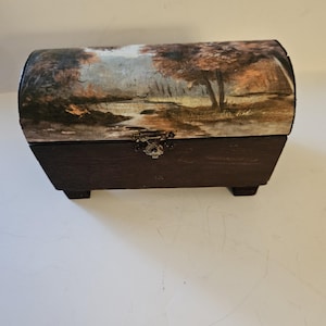 May include: A small, wooden chest with a curved lid. The lid features a landscape painting with trees and a river. The chest's base is dark brown, and it has a decorative metal clasp. The chest is elevated on small feet.