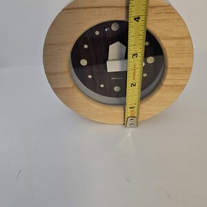 Puede incluir: Un reloj redondo de madera con una esfera oscura y un dise&ntilde;o plateado en forma de casa. El reloj tiene aproximadamente 10 cm de di&aacute;metro. Tiene un marco de madera clara y una cubierta de pl&aacute;stico transparente.
