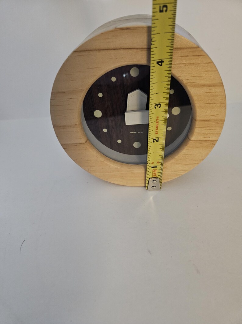 Puede incluir: Un reloj redondo de madera con una esfera oscura y un dise&ntilde;o plateado en forma de casa. El reloj tiene aproximadamente 10 cm de di&aacute;metro. Tiene un marco de madera clara y una cubierta de pl&aacute;stico transparente.