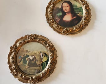 Puede incluir: Dos pequeñas impresiones redondas enmarcadas de color dorado. Una presenta la pintura de la Mona Lisa, la otra representa una escena de personas trabajando en un campo. Los marcos tienen detalles ornamentados y arremolinados.