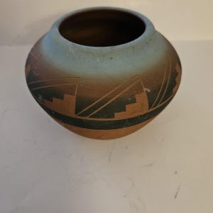 May include: A round, handcrafted pottery vase with a wide opening. The vase features a gradient of colors, transitioning from a light blue at the top to a brown base. Geometric black designs encircle the middle, adding a touch of artistry to the ceramic piece.
