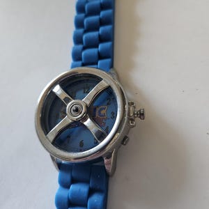 Puede incluir: Un reloj de pulsera azul con bisel plateado y correa de silicona azul. La esfera del reloj presenta el texto "Sonic" y un dise&ntilde;o en forma de cruz. La correa est&aacute; hecha de eslabones.