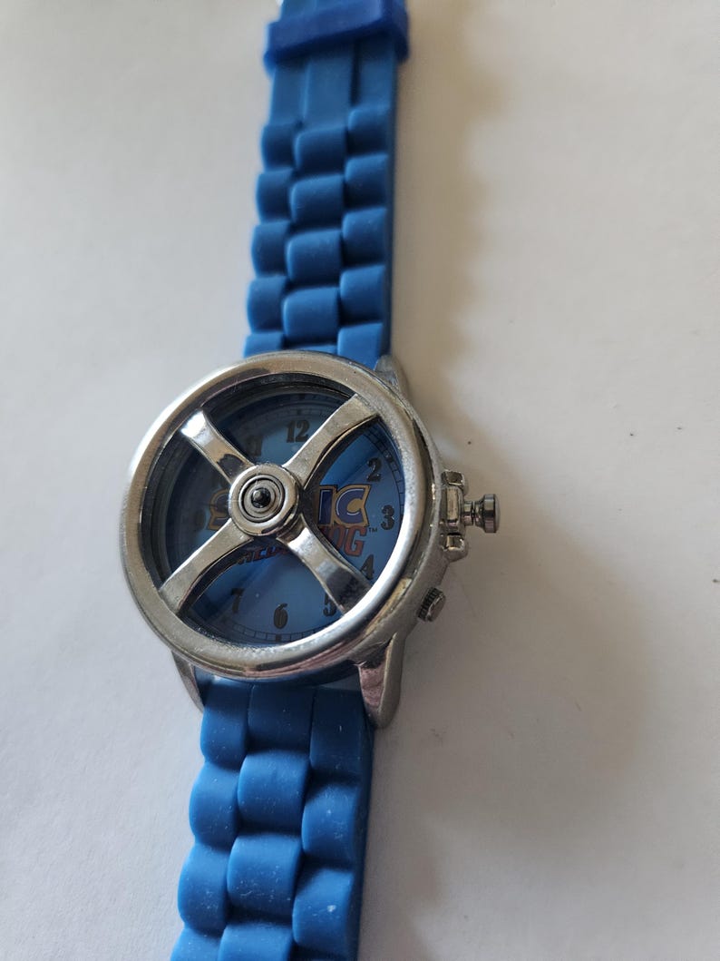 Puede incluir: Un reloj de pulsera azul con bisel plateado y correa de silicona azul. La esfera del reloj presenta el texto "Sonic" y un dise&ntilde;o en forma de cruz. La correa est&aacute; hecha de eslabones.