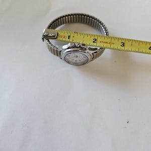 May include: A silver-toned wristwatch with a white face and black numerals. The watch has a stretch band and is measured with a yellow tape measure. The watch band measures approximately 1 inch in diameter.
