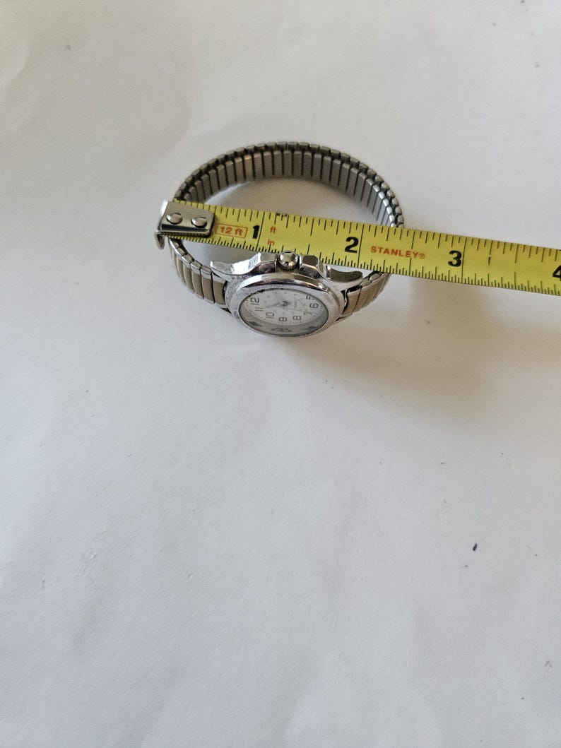 May include: A silver-toned wristwatch with a white face and black numerals. The watch has a stretch band and is measured with a yellow tape measure. The watch band measures approximately 1 inch in diameter.
