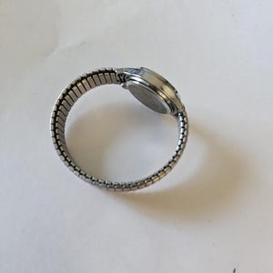 May include: A silver-toned wristwatch with an expandable band. The watch face is round and the band is made of small, connected metal links. The watch is shown on a white surface.