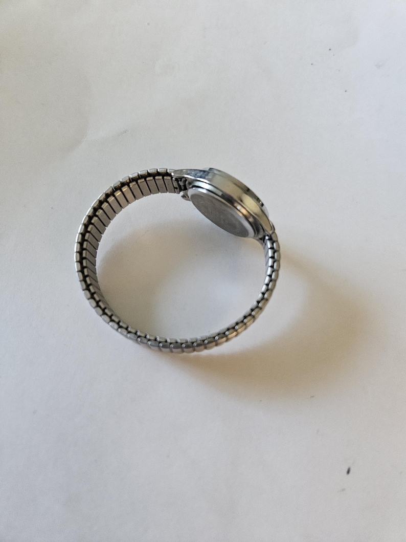 May include: A silver-toned wristwatch with an expandable band. The watch face is round and the band is made of small, connected metal links. The watch is shown on a white surface.