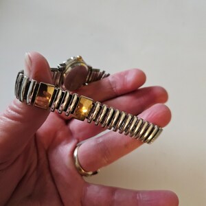 May include: A silver and gold tone watch with a metal band. The watch has a round face and a gold tone bezel.